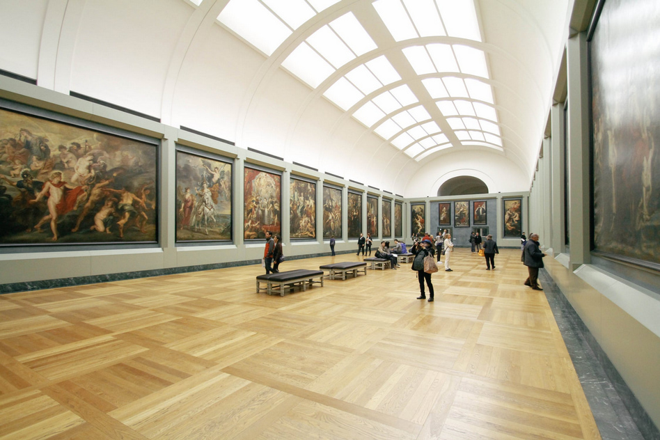 A spacious art gallery with an arched ceiling and a series of large, classical paintings displayed in gold frames on the walls. 